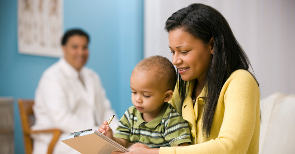 A woman in a yellow sweater helps a young boy with a pen and a notebook while a doctor observes in the background